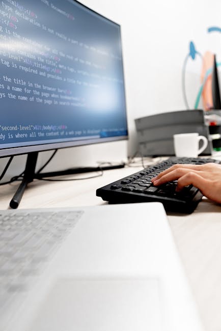 A person operates a keyboard in front of a computer monitor displaying lines of programming code, situated on a tidy desk. The workspace includes a white keyboard, a gray laptop, and various office su