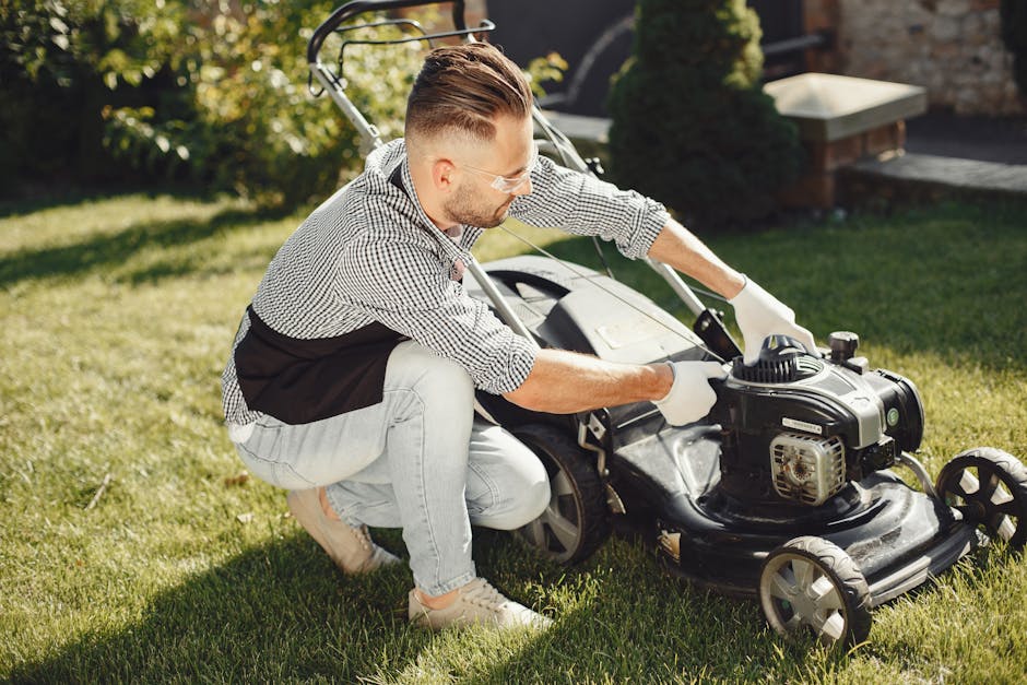 A man wearing glasses, a checkered shirt, and light-colored jeans is kneeling on a neatly trimmed lawn, operating a petrol-powered lawn mower for garden maintenance. The lawn mower has a black and sil